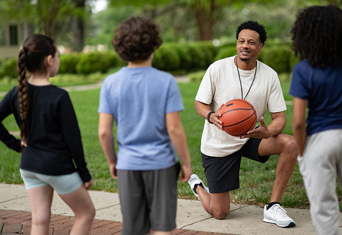 Warrior kneels outdoors holding a basketball while coaching a group of children during a community activity.