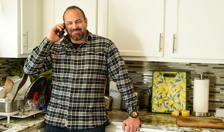 Warrior Benjamin Heck in a plaid flannel shirt stands in a kitchen while talking on a cellphone, leaning against a countertop with white cabinets.