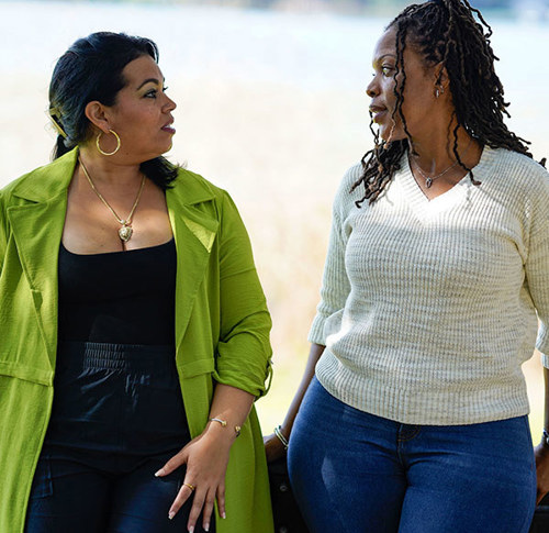 Two women walk side by side, engaged in conversation. One wears a bright green coat, the other a white sweater. They look at each other thoughtfully.