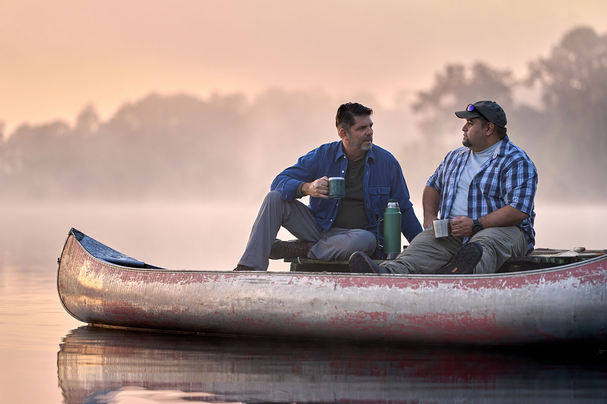 Wounded Warrior Tim Aponte sits with a fellow veteran on a lake dock with a canoe, drinking coffee and smiling.