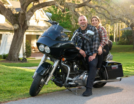 Wounded Warrior Bill Geiger and his daughter sitting on motorcycle parked on a residential street, surrounded by greenery with sunlight filtering through the branches.
