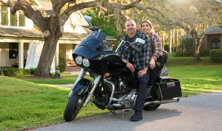 El veterano herido Bill Geiger y su hija sentados en una motocicleta estacionados en una calle residencial, rodeados de vegetación con la luz del sol filtrándose a través de las ramas.
