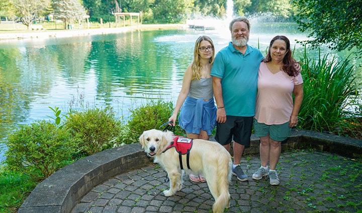 Warrior David Denhardt, his family, and a service dog stand on a stone-paved path beside a pond with a fountain, surrounded by trees and greenery.