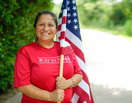 La veterana herida Maria Edwards usa una camiseta roja de WWP Fit mientras sonríe y sostiene una bandera estadounidense a lo largo de un camino arbolado.
