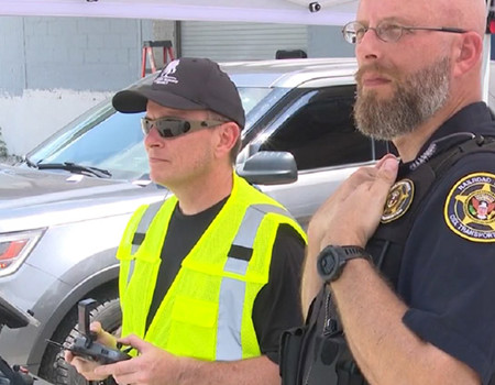 A wounded warrior wearing a yellow safety vest and sunglasses is operating the controls of a drone while a CSX police officer stands by him providing instruction.