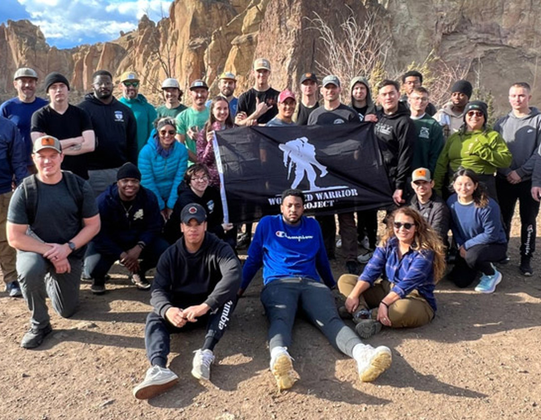 A group of active-duty service members pose together holding a Wounded Warrior Project flag after hiking.