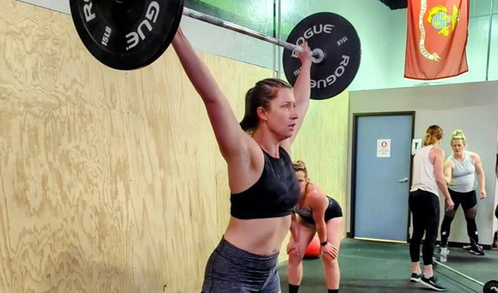 Una mujer levanta una barra por encima de la cabeza en un gimnasio de CrossFit durante la recaudación de fondos del desafío Murph, rodeada de otros participantes.