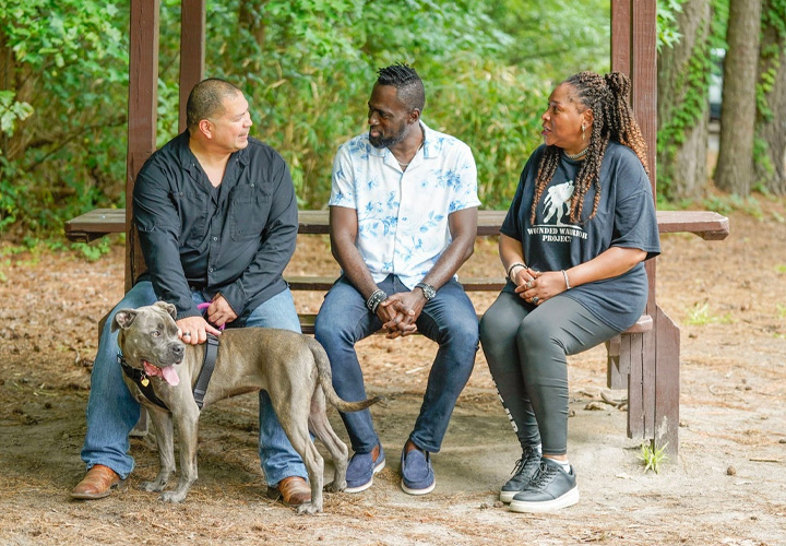 Wounded warriors Eric Frank, Adams Amisu, and Barbara Reese sit on a bench alongside a nature trail with Eric’s service dog. They are deep in conversation.