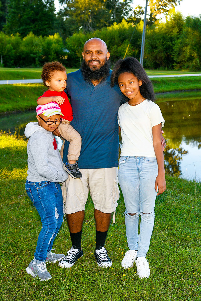Wounded warrior Eric Delion stands smiling with his three children next to a pond. A white pathway bridge and trees are off in the distance.