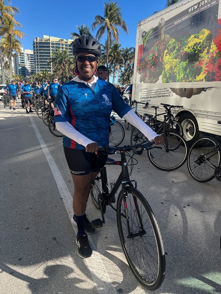 Jackie Gordon stands with her bike on a palm-lined street with other warriors behind her.