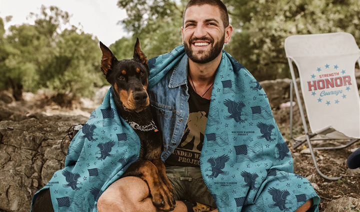 Man holding a Doberman dog with a teal blanket wrapped around his shoulders, smiling. A camping chair is in the background.