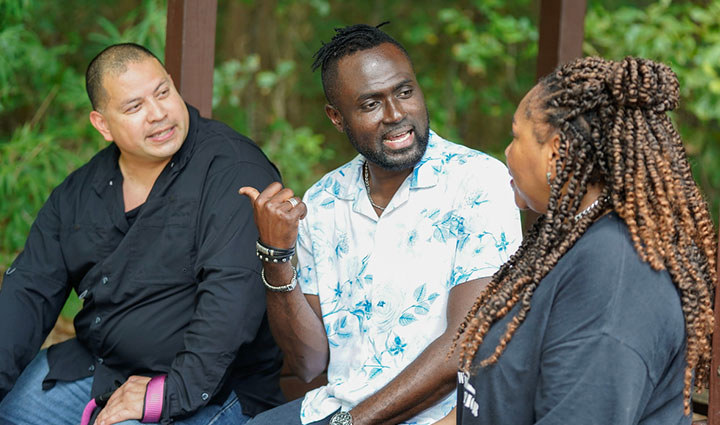 Warriors Eric Frank, Adams Amisu, and Barbara Reese sit and talk together on a bench in an outdoor pavilion, surrounded by green trees and foliage.