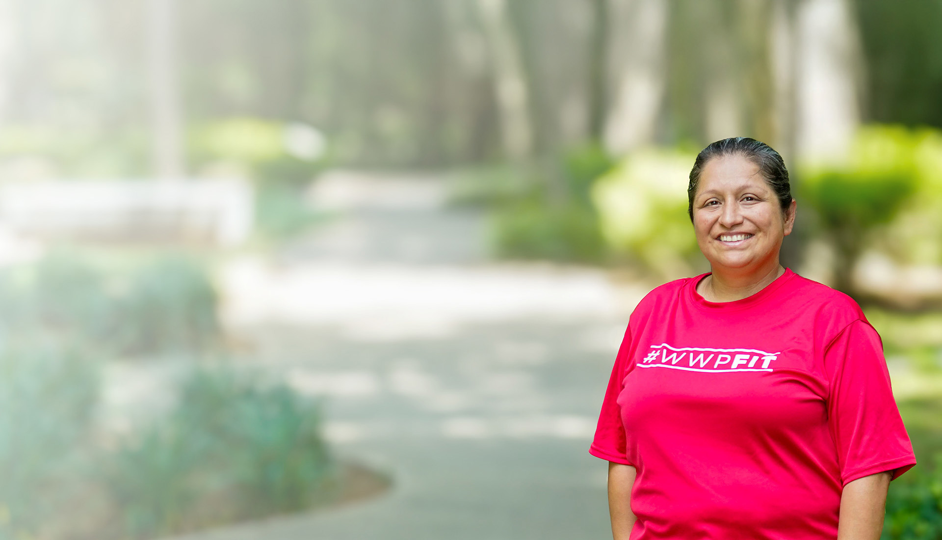 Wounded warrior Maria Edwards stands smiling on a outdoor pathway surrounded by greenery. She is wearing a bright red 
