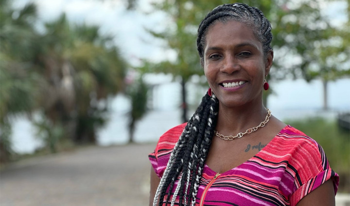 La veterana Tonya Oxendine lleva una blusa de colores vivos rosa, violeta y amarillo y su larga trenza cae sobre su hombro, con un fondo de cielo azul y palmeras verdes.