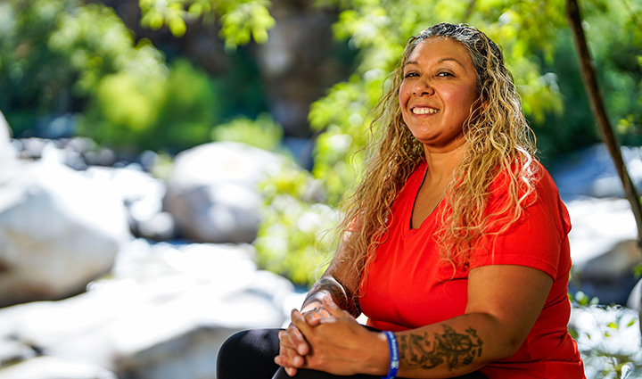 Surrounded by nature, wounded warrior Ysabel Cardona sits on a large rock in Sequoia National Park, smiling at the camera.