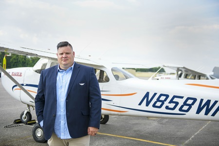 Jacob Norotsky in a blazer and button-down shirt stands in front of a small airplane on a tarmac.