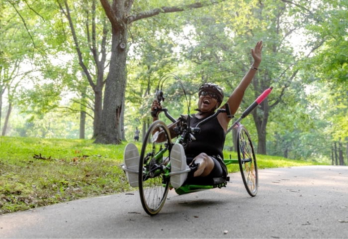 Wounded warrior Donna rides a recumbent bike on a path through a park, raising her arm in joy and accomplishment.