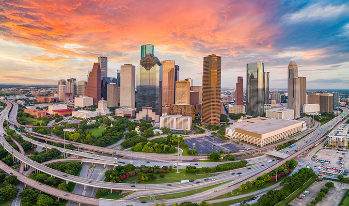 Aerial view of downtown Houston at sunset with skyscrapers, highways, green spaces, and a sky glowing orange, pink, and blue. 