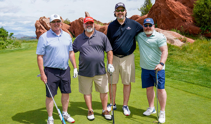 Four men, dressed in golf attire, stand side by side on a golf course with a clear blue sky and red rock formations in the background.