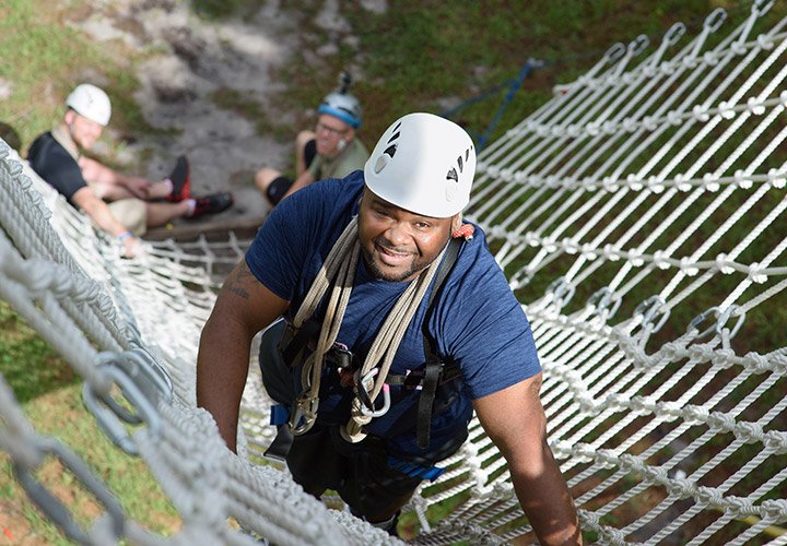 Equipped with a harness and helmet, wounded warrior Chris Gordon ascends a net, making his way toward the ropes course platform.