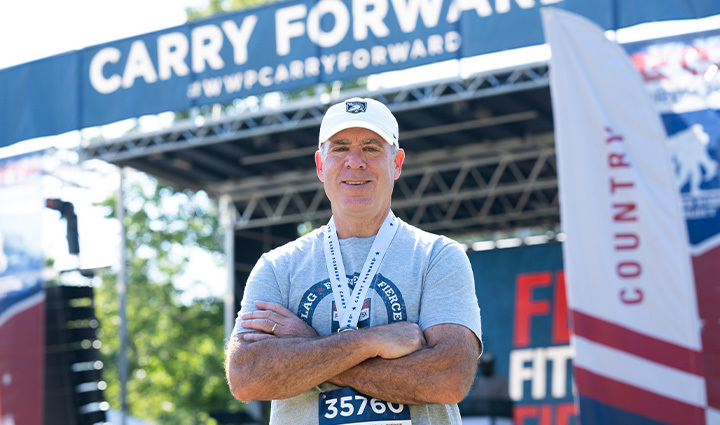 A WWP Carry Forward 5K participant, Doug Frank, standing in front of a Carry Forward 5K stage with a medal around his neck. 