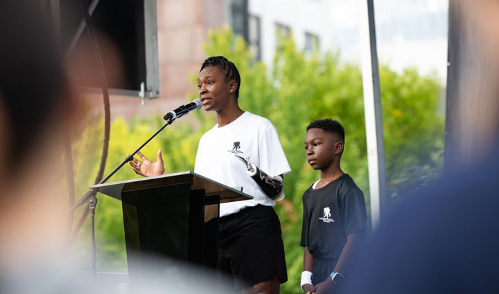 Wounded Warrior Danielle Green stands at a podium and speaks into a microphone as her son stands behind her.