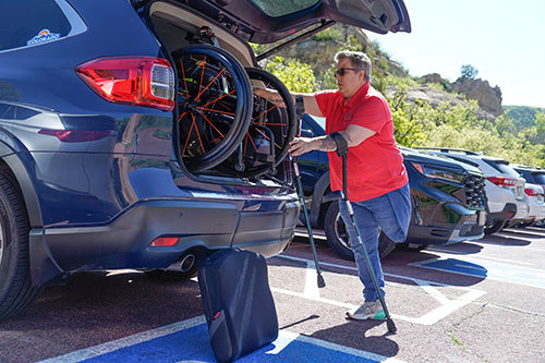 Carla stands with crutches, looking at her wheelchair in the trunk of a car.