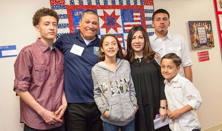 A wounded warrior stands in front of a patriotic quilt and smiles alongside his family. 