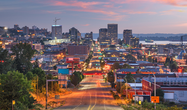 Downtown Tacoma at dusk with lit buildings, Puget Sound in the background, and a sky glowing in blue and pink sunset hues.