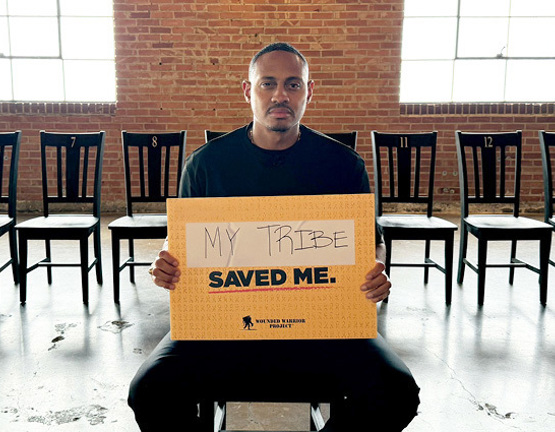 Warrior Tyshawn Jenkins sits in a chair in front of a line of chairs, holding a sign that says 