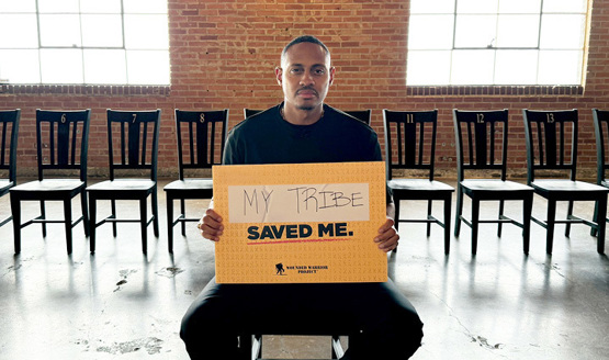 Warrior Tyshawn Jenkins sits in a chair in front of a line of chairs, holding a sign that says 