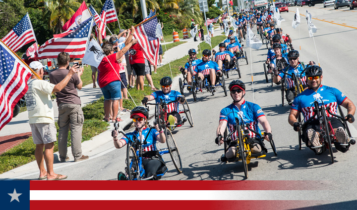 A group of warriors riding recumbent bicycles as a crowd with American flags cheers them on from the sidewalk.