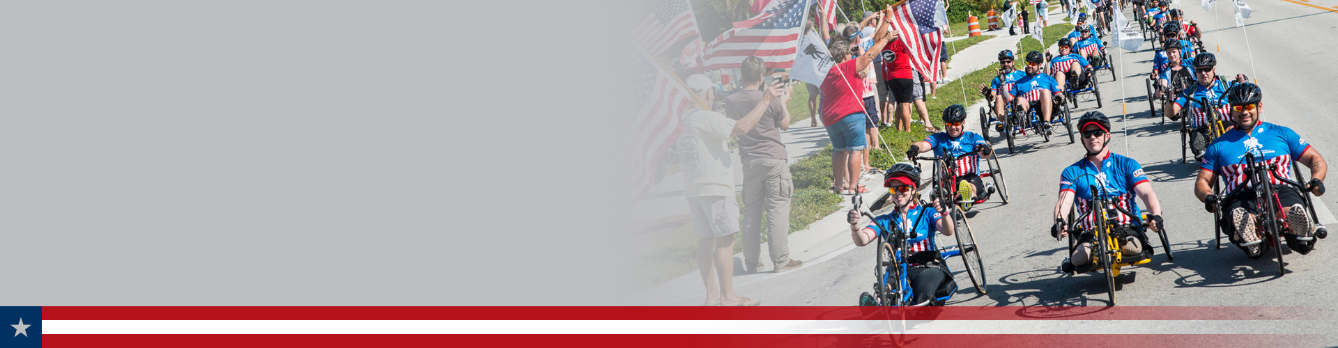 A group of warriors riding recumbent bicycles as a crowd with American flags cheers them on from the sidewalk.