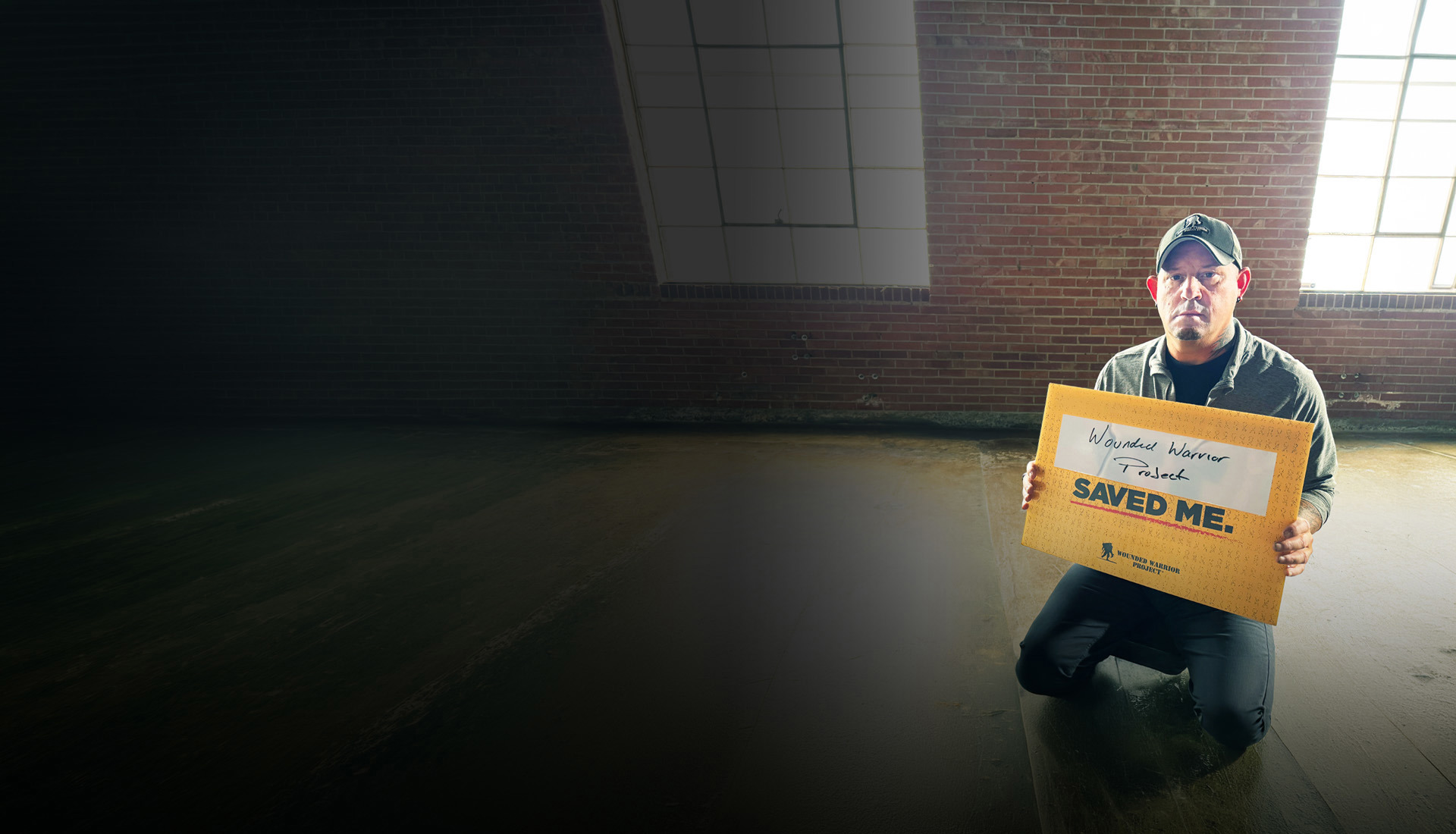 Warrior Phil Krabbe sits in front of a brick wall, holding a yellow sign that says, 
