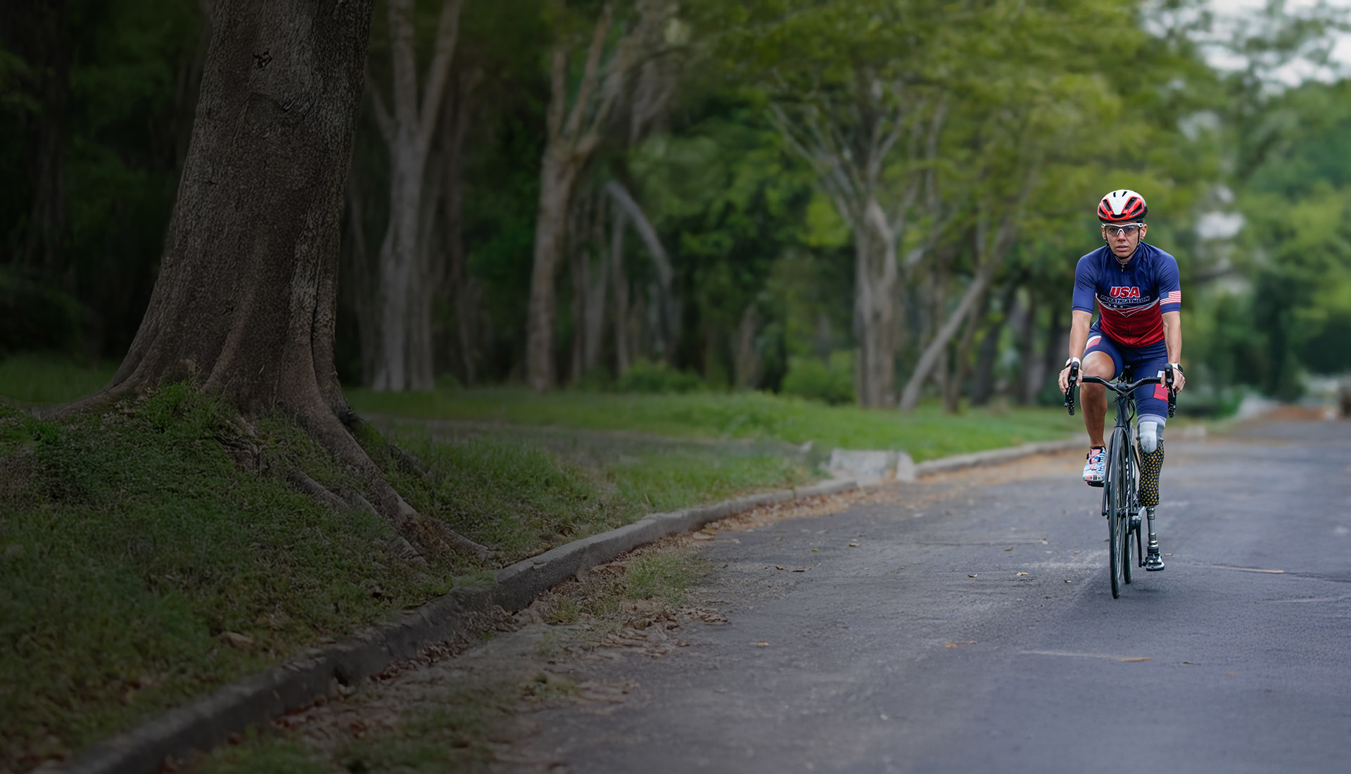 Warrior Kelly rides a road bike on a tree-lined street, wearing a USA cycling kit and helmet, with a prosthetic leg.