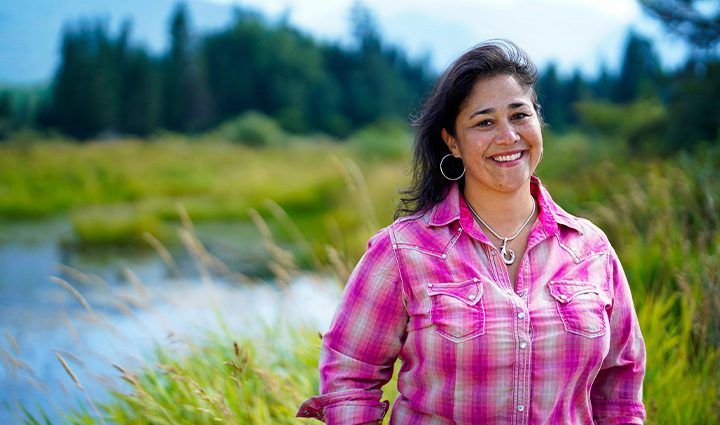 Wounded warrior Tina Waggener stands in front of a scenic background of mountains, trees, and a stream, smiling at the camera. 