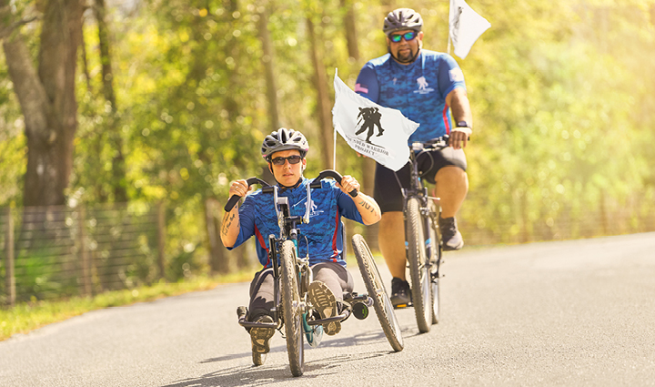 Warriors Beth King and Tim Aponte ride adaptive cycles on a paved road, each carrying a flag, surrounded by trees. 