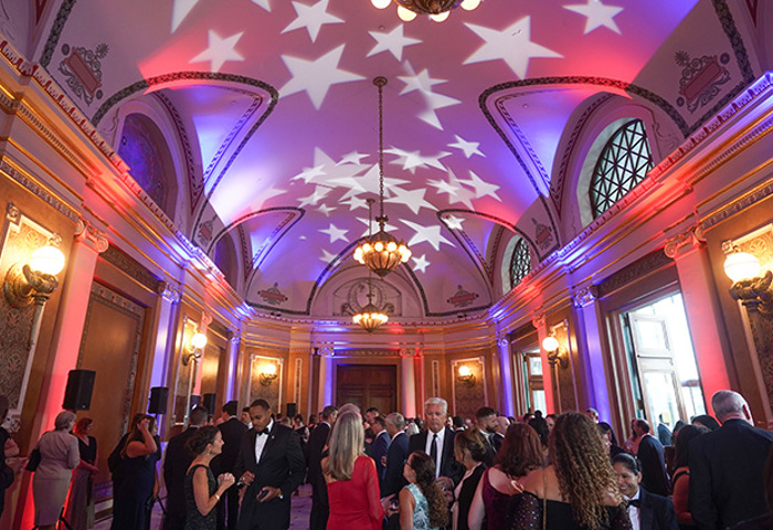 A grand ballroom with the celling illuminated with red, white, and blue lights projecting star shapes. The room is filled with people dressed in formal attire, engaging in conversation and socializing for WWP's Courage Awards and Benefit Dinner.