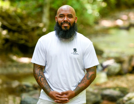 Wounded warrior Eric Delion stands in front of a stony brook surrounded by trees while clasping his hands and smiling at the camera.