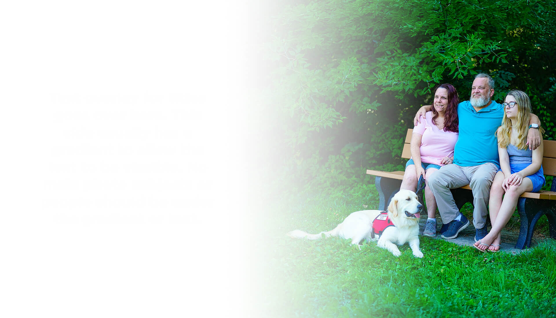 Wounded warrior David Denhardt and his family sit together on a park bench, smiling and surrounded by greenery, with his service dog lying in the grass in front of them.