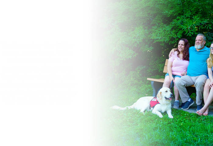 Wounded warrior David Denhardt and his family sit together on a park bench, smiling and surrounded by greenery, with his service dog lying in the grass in front of them.