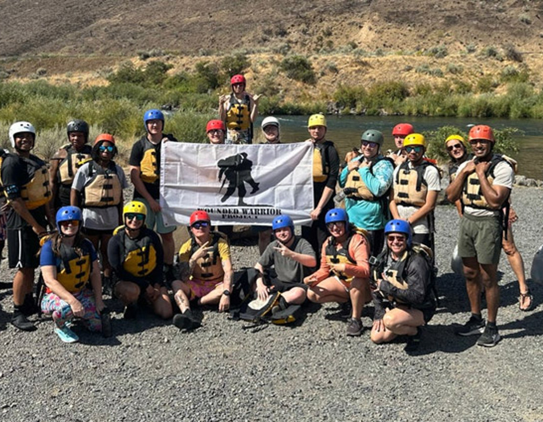 A group of active-duty service members pose together holding a Wounded Warrior Project flag after whitewater rafting.