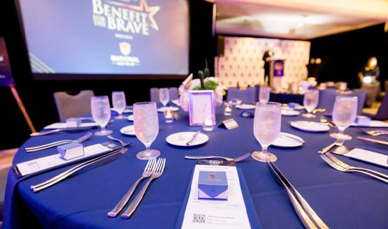 Mesa de Benefit for the Brave con mantel azul, menús, vasos y cubiertos. Hay una pantalla con una presentación en el fondo.