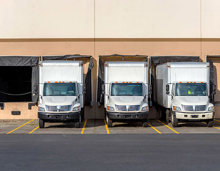 Three semi-trucks with box trailers parked in row at a warehouse.