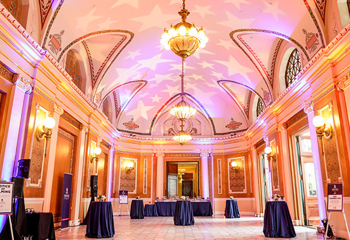 Elegant event hall with high arched ceilings, ornate chandeliers, and warm lighting, set with cocktail tables.