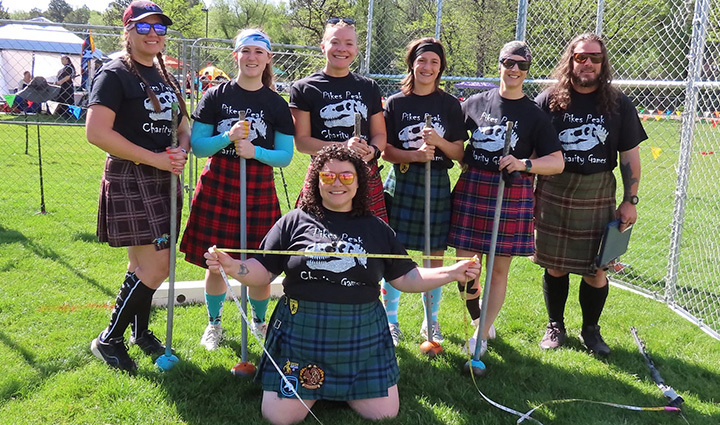 Un grupo de siete personas, vestidas con faldas escocesas y camisetas negras, posan con herramientas pesadas para los Pike's Peak Charity Games en un campo de césped.