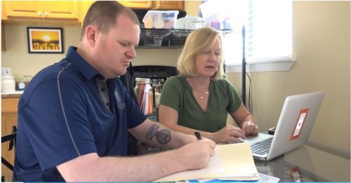 A wounded male warrior sitting at a table looking at a computer with a caregiver.