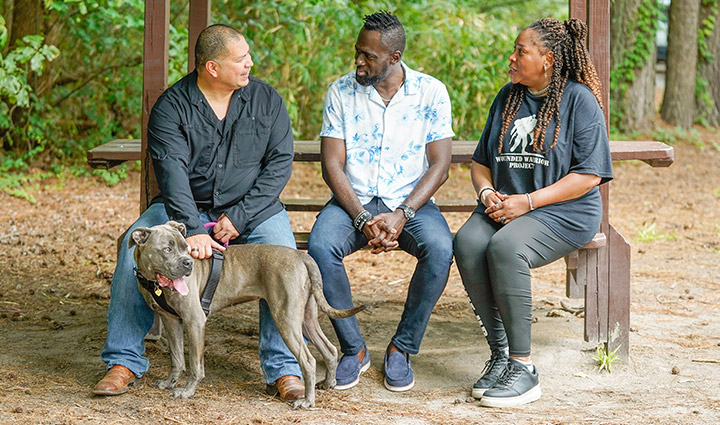 Warriors Eric Frank, Adams Amisu, and Barbara Reese all sit on a bench outside talking together.