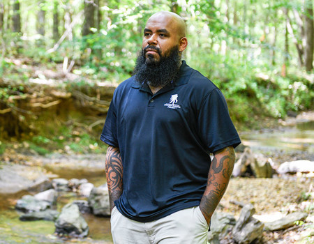 Wounded warrior Eric Delion stands in front of a stony brook surrounded by trees, appearing to be deep in thought while his hands are in his pockets.