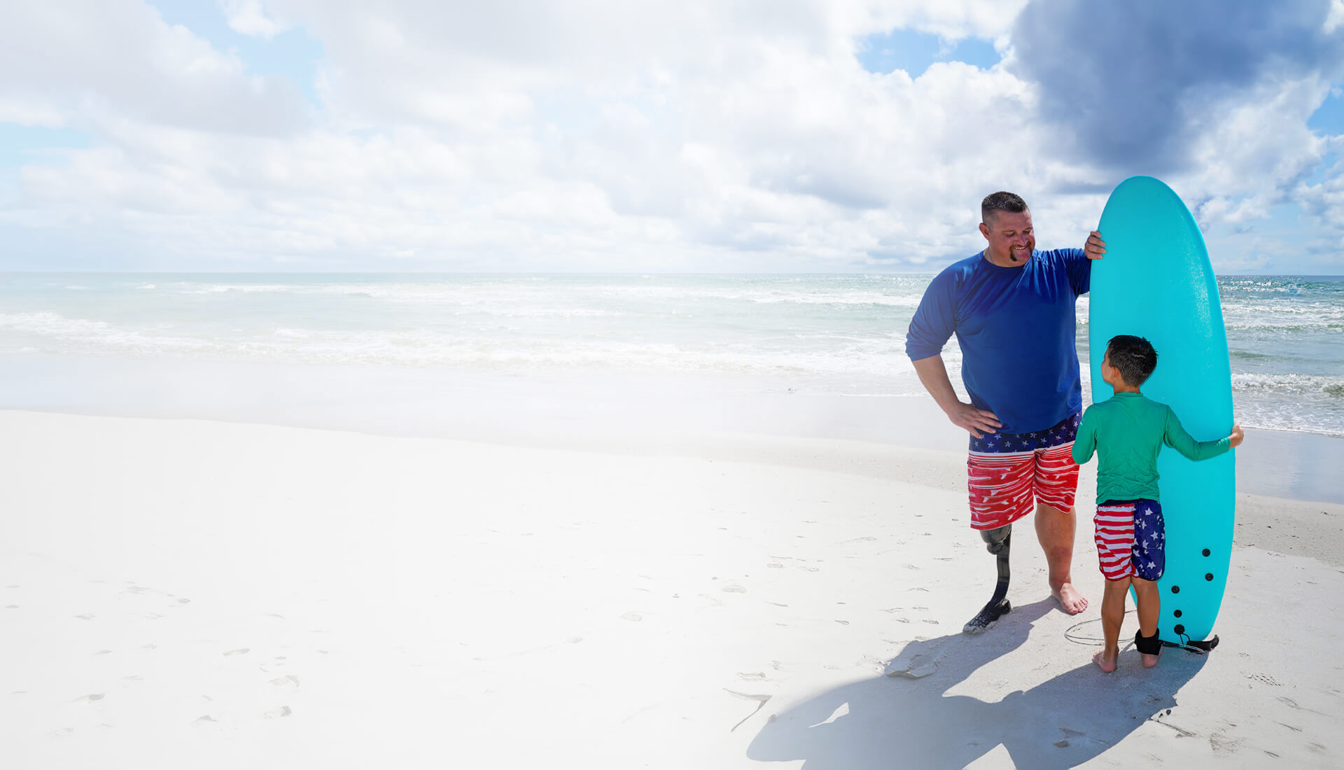 Wounded warrior Bryan Wagner with a prosthetic leg and his young son stand on a beach, holding a surf board together.